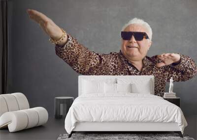 Retired senior man vibing to hype pop music. Portrait of funny rich white haired old grandpa in cool glasses and leopard patterned party shirt flexing, dancing and having fun on gray studio background Wall mural