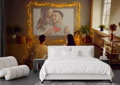 Romantic couple having Valentine's Day date at home, using modern projector in living room interior with beautiful fairy lights, watching movie, looking at digital photos, enjoying memories. Back view Wall mural