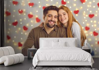 Saint Valentine's Day celebration, gift giving and romantic couple concept. Portrait of happy smiling young man and woman hugging and holding red present box against lights and hearts decor background Wall mural