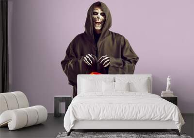 Scary man with skull makeup in hooded black cloak. Creepy man wearing Halloween costume holding scary pumpkin lantern standing on light lilac background. Halloween party concept Wall mural