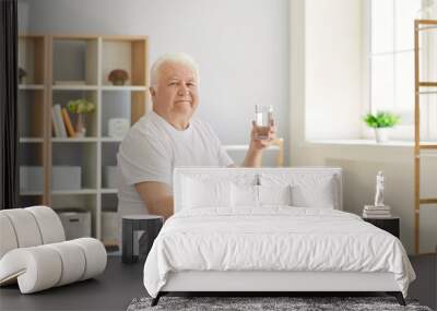 Senior man holding a glass of water sitting on a fitball at home and looking at the camera. Concept of active lifestyle and maintaining water balance in the elderly. Wall mural