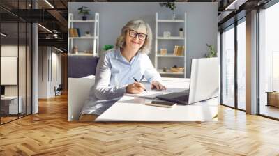 Senior smiling woman sitting on workplace at the desk working on laptop making notes planning budget. Concept of e-learning for mature people or distance freelance work career for retirement. Wall mural