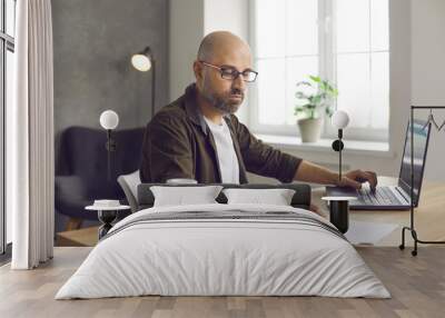 Serious bald business man in glasses sitting at desk in home office, working on laptop computer, searching for information, taking notes, writing down important details, drinking coffee from paper cup Wall mural