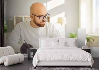 Serious man in glasses and comfortable fingerless compression hand gloves for easing arthritis pain sitting at desk, using computer, typing on keyboard, holding pen and taking study notes on paper Wall mural