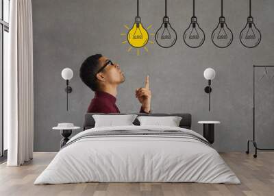 Side profile view young man making choice looking up at several electric light bulbs. Creative Afro American entrepreneur considers different effective business ideas, points finger and picks best one Wall mural