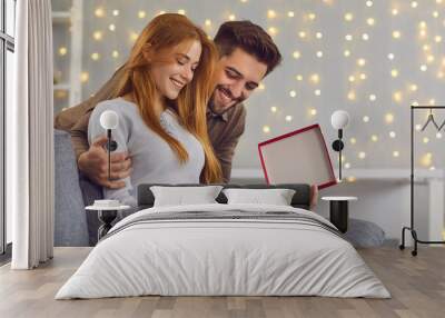 Side view of a red-haired young woman with her husband opening a gift box while sitting at home on the couch. Couple in love looks contentedly at the contents of the box. Valentine's day concept. Wall mural