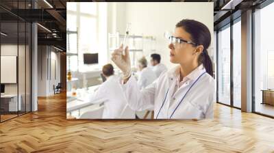 Side view young female scientist in coat, gloves and goggles looking at small green leaf inside test glass tube in her hand. Nature and science, using plants in cosmetology laboratory research concept Wall mural