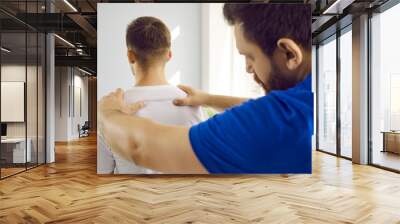 Skilled male therapist examining young man's back and appoints physiotherapeutic treatment. Close up of orthopedist examining man's back in modern health clinic. Health care concept. Wall mural