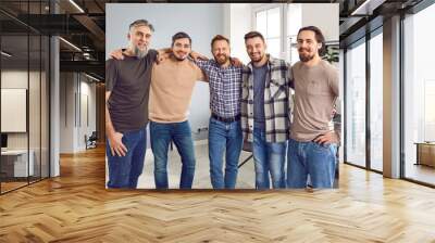 Smiling five male friends standing and hugging shoulders together. Portrait of positive cheerful hipster men wearing casual clothes smiling at camera indoors. Male friendship concept Wall mural