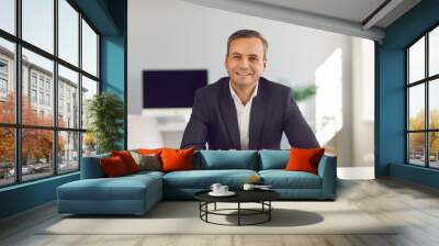 Smiling middle aged businessman in suit sitting and looking at camera during online videocall or interview with candidate in office over modern company interior. Working in office, online business Wall mural