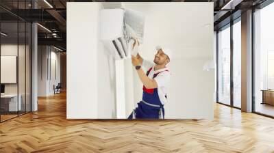 Smiling technician in new beautiful uniform changes air conditioner filter element in apartment. Handsome worker cleaning ventilation system indoors. Warranty service for household appliances. Wall mural