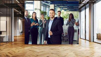 Smiling young attractive boss and business leader man proudly looking at camera with team of people applauding in background in office. Business team or group of staff congratulating their leader. Wall mural