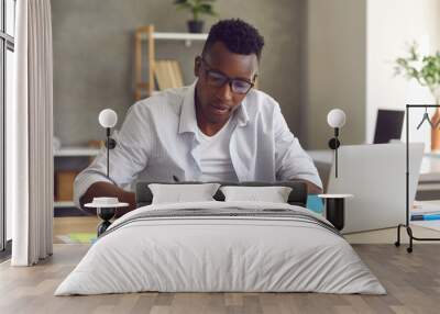 Startup project development, research work, brainstorm. Young handsome minded black african american businessman sitting at home table front of laptop writing down ideas in notebook Wall mural