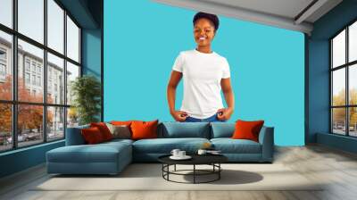 Studio portrait of happy woman in casual clothes. Beautiful confident young African American girl in blue jeans and white mockup T shirt standing isolated on turquoise background. Fashion concept Wall mural