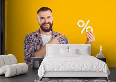 Studio portrait of smiling man with percentage sign. Happy young man in casual shirt isolated on orange yellow background advertising sale and pointing at discount percent symbol in his hand Wall mural