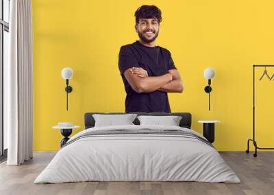 Studio shot of happy young man in casual wear. Handsome Indian guy with modern haircut in black Tshirt standing with his arms folded isolated on yellow background, looking at camera and smiling Wall mural