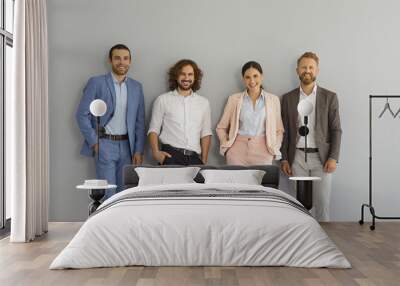 Successful stylish business team standing in studio. Group portrait of four happy confident young people in smart office suits standing together against grey wall, looking at camera and smiling Wall mural