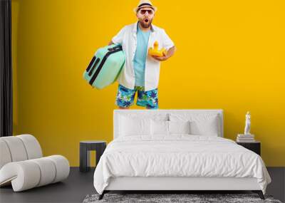 Surprised summer tourist. Full length bearded man in white blue holiday wear, sun hat and sunglasses holding toy and green travel bag standing with amazed face expression on yellow orange background Wall mural