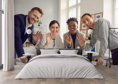 Team of happy young diverse business people sitting and standing around office table, smiling, looking at camera and waving hello, greeting international coworkers in remote work meeting via Internet Wall mural