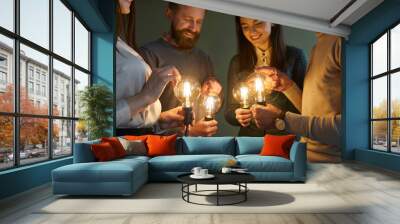 Team of people developing and trying out innovative ideas. Group of four clever men and women holding bright, shining, glowing Edison light bulbs to illuminate concept of creative idea and innovation Wall mural