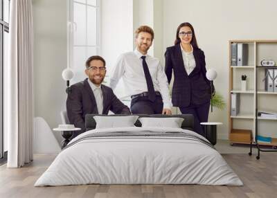 Team of three young business people or corporate employees in office. Group portrait of two happy men and one woman in formal wear standing by desk or sitting on chair, looking at camera and smiling Wall mural