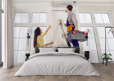 Technician repairing AC on his typical working day. Man climbs ladder with toolbox in order to check, do disinfection or fix troubles in modern white wall mounted air conditioner in young lady's home Wall mural