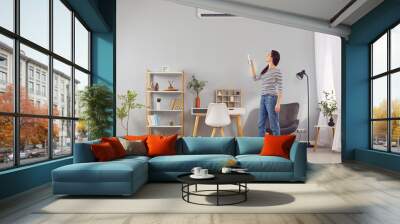 Tenant or apartment owner switches modern air conditioning system at home. Young woman standing in beautiful living room interior and setting up temperature on air conditioner on gray wall. AC concept Wall mural