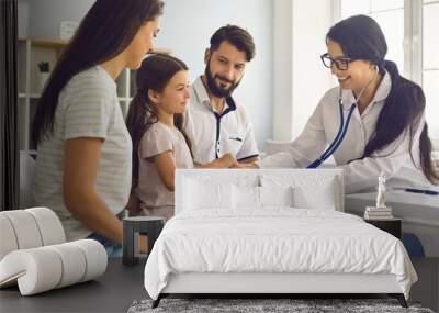 The family doctor listens with a stethoscope to the child with the parents sitting at the table in the clinic. Wall mural