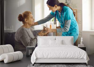 Treatment, care and support. Friendly young doctor visiting female patient at home. Caring nurse at clinic or assisted living facility helping senior woman take medicine and giving her glass of water Wall mural