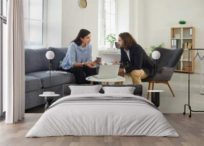 Two business people meeting and having discussion in modern office interior. Young man and woman sitting on chair and sofa by table with laptop, drinking coffee and talking about work Wall mural