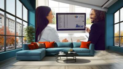 Two cheerful colleagues in office are discussing joint work project and analyzing financial graphs. Man and woman look at each other and smile while sitting in front of working computer monitor. Wall mural