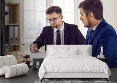 Two professional male lawyers read legal documents and study information about court case together. Men in suits sit in office at table near judge gavel and scales of justice and discuss legal issues Wall mural