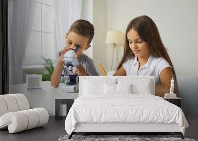 Two school children use microscope and look at microorganisms at magnification. Little boy studying something under microscope while little girl is taking notes on notepad for scientific hobby project Wall mural