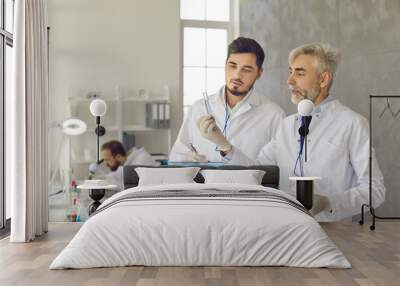 Two scientists in white lab coats and gloves developing new effective drug together. Serious young student trainee with clipboard making notes and talking to senior professor in university laboratory Wall mural