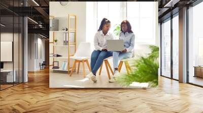 Two young African American women sitting on chairs in modern office interior, using laptop computer together, working on business project, and doing online research Wall mural