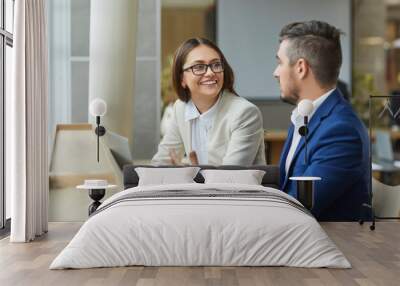 Two young smiling company employees man and woman sitting with laptop having discussion, talking about new job projects or startups. Business people working together in modern office. Wall mural