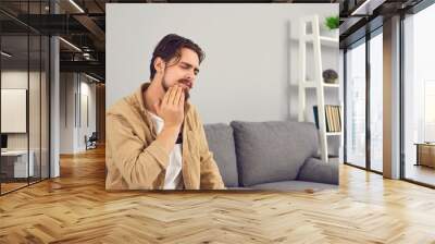 Upset man suffering from severe toothache and touching cheek with grimace of pain. Young guy suffering from very sensitive teeth or inflamed tooth nerve. Concept of people having dental problems Wall mural