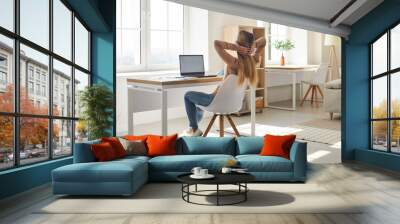 Woman feeling happy after work is done. College or university student girl holding hands behind head and looking out window while relaxing in comfy chair by desk with modern laptop computer at home Wall mural