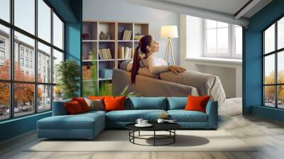 Woman uses chance to relax, unwind and get some peace of mind on comfy couch at home. Happy lady enjoying silence and quiet leisure time in beautiful room with comfortable sofa and Nordic wooden shelf Wall mural