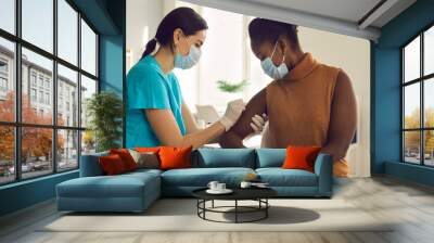 Young African American woman getting flu shot during seasonal vaccination campaign. Doctor or nurse in medical face mask cleans skin on patient's arm before injecting modern Covid 19 antiviral vaccine Wall mural