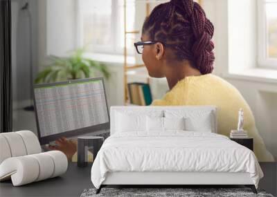 Young African American woman with Afro braids sitting at her desk at home or in the office, working on her laptop computer and using modern business spreadsheets. View over the shoulder Wall mural