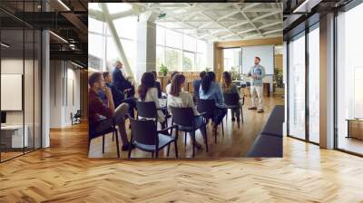 Young berded man speaking on a business training standing in office during a conference or seminar for a group of diverse people company employees sitting on chairs in meeting room. Wall mural