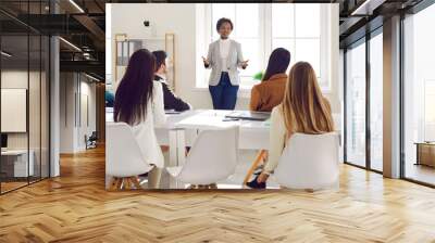 Young business people sitting around big table and listening to team leader. Black woman standing in modern white office boardroom during meeting and speaking to group of multiracial colleagues Wall mural