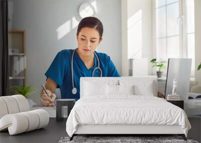 Young busy female nurse is concentrating on working while sitting at desk at workplace in clinic. Girl with stethoscope around her neck works at laptop and makes notes in paper documents. Wall mural