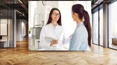 Young cheerful Caucasian woman doctor and female patient talking in modern cosmetology clinic, beautician in white coat smiling consults girl in shirt stands in bright room for SPA procedures Wall mural