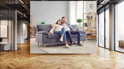 Young family couple spending time at home. Happy, relaxed man and woman sitting together on a comfortable sofa in a modern living room interior in their house or apartment Wall mural