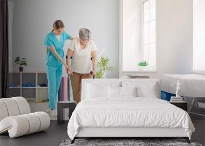 Young female caregiver, nurse helping elderly woman to walk with crutches during rehabilitation in clinic. Physiotherapist providing assistance and supporting senior patient after injury or stroke. Wall mural