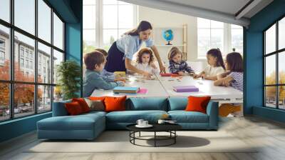 Young female caucasian teacher working with little children group in classroom explaining. Woman tutor and schoolkids joining puzzle. Preschool or primary school education and development Wall mural