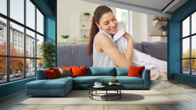 Young housewife smells clean clothes. Happy, smiling woman sitting on the couch with a laundry basket, holding a clean, fresh, white shirt and smelling a nice, pleasant smell of the fabric conditioner Wall mural