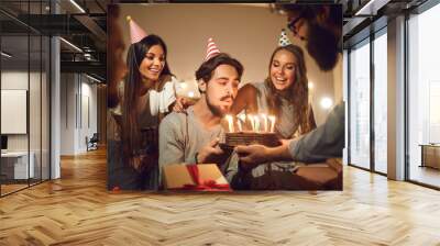 Young man blowing candles on his birthday cake surrounded by happy smiling friends Wall mural